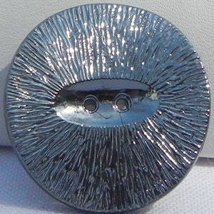 May include: A large, round, silver button with a textured, radial design. The button has an oval-shaped center with two holes and a small, curved line, resembling a smiling face. The button is likely for clothing or crafts.