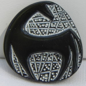 以下が含まれることがあります： スタイリッシュなデザインの黒い楕円形のボタン。ボタンは、盛り上がった銀色の幾何学模様と滑らかで光沢のある仕上がりを特徴としています。デザインは、動物または人物の抽象的な表現のようです。