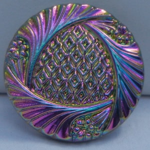 Peut inclure: Un bouton rond et irisé avec un motif central en losanges et des bords tourbillonnants texturés. Les couleurs du bouton varient du violet au vert et au bleu, avec de petits accents floraux.