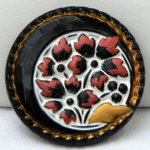 May include: A round, black button with a floral design. The center features a white background with red and black leaves and black dots. The outer edge is black with a gold trim.