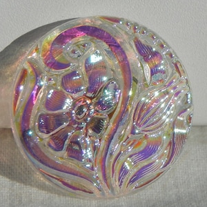 Könnte beinhalten: Ein klarer, irisierender Glas-Knopf mit floralem Design. Der Knopf zeigt eine zentrale Blume und wirbelnde Blattmuster, mit Farben, die von Lila nach Gelb wechseln. Der Knopf ist rund und hat eine detaillierte, geprägte Oberfläche.