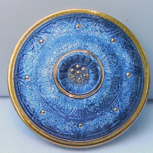 May include: A round, blue and gold decorative button. The button features a detailed, textured blue surface with a floral design and gold accents. The outer rim is gold, and the center has a smaller gold circle with gold dots.