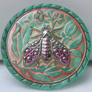May include: A round, decorative button with a detailed insect design. The button features a silver-colored insect with pink accents, surrounded by green leaves and a green border. The background has a peach and cream color scheme.