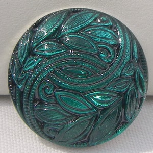May include: A round, teal-colored button with a detailed leaf and vine design. The button has a glossy finish and a raised, textured pattern. The design is symmetrical and features swirling elements.
