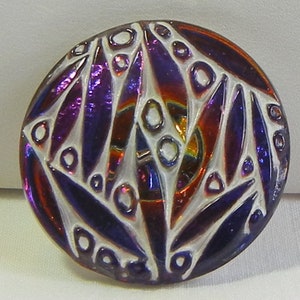 May include: A round button with a white, raised design and iridescent purple, red, and blue segments. The button has a glossy finish and a decorative pattern, suitable for sewing or crafts.