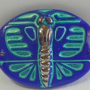 May include: An oval-shaped, blue decorative object with a butterfly design. The butterfly has silver-colored body segments and teal-colored wings. Two small, silver-colored studs are on either side of the oval. The object has a glossy finish.