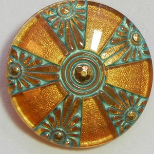 May include: A round, decorative button with a textured, golden-orange surface. The button features teal-colored accents and a central design with radiating segments and floral-like details. Small, gold-colored embellishments are placed throughout the design.