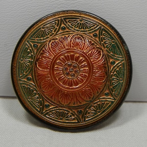 May include: A round, decorative button with an embossed floral design. The button features a central orange flower surrounded by green and gold accents. The button has a dark brown border.