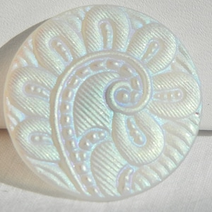 May include: A round, iridescent white button with a floral design. The button features a spiral center and petal-like shapes with textured lines and small, raised dots. The button has a pearlescent sheen.