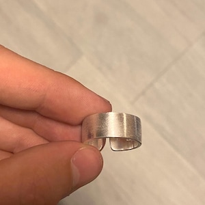 Könnte beinhalten: Ein breiter silberfarbener Ring mit gebürstetem, mattem Finish und einer sichtbaren Öffnung an der Unterseite. Das Band hat eine flache, gleichmäßige Oberflächenstruktur, die das Licht gleichmäßig auf der metallischen Außenseite reflektiert.
