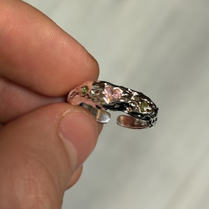 Könnte beinhalten: Ein verstellbarer Ring in Silber mit strukturiertem Band. Der Ring ist mit kleinen, herzförmigen rosa und runden grünen Edelsteinen besetzt. Der Ring wird von den Fingern einer Person gehalten.