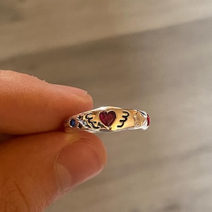 Könnte beinhalten: Ein silberfarbener Ring mit einem zentralen herzförmigen roten Edelstein, flankiert von schwarzen, gravierten Wellenlinien und kleinen Sternmotiven. Das Band enthält einen kleinen blauen runden Stein auf der einen und einen roten Stein auf der anderen Seite.