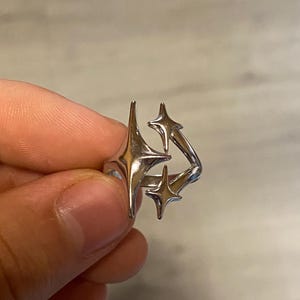 Könnte beinhalten: Ein silberfarbener Metallring mit einer Anordnung aus drei vierzackigen Sternen in unterschiedlichen Größen. Die Sternmotive sind asymmetrisch entlang der Ringschiene platziert und weisen eine polierte, reflektierende Oberfläche auf.