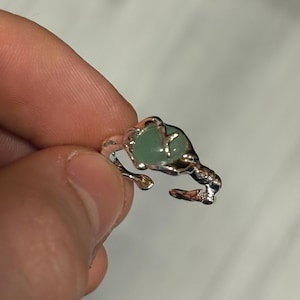 Könnte beinhalten: Silberfarbener Ring mit unregelmäßigem, organischem Design. Der Ring hat einen hellgrünen, oval geformten Stein in der Mitte. Das Band hat ein strukturiertes, astähnliches Aussehen und erzeugt eine einzigartige Ästhetik.