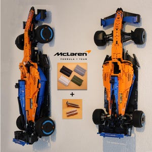 Könnte beinhalten: Ein Lego McLaren Formel 1 Team Rennwagenmodell in Orange und Blau mit schwarzen Rädern. Das Auto ist mit Schrauben an einer Wand befestigt. Das Auto wird mit einem McLaren-Logo und den Worten "Formel 1 Team" präsentiert.