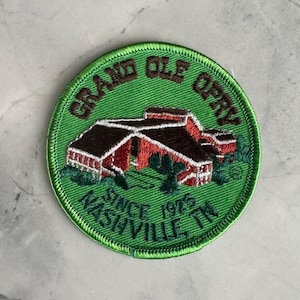 May include: Circular embroidered patch with a green border, featuring the Grand Ole Opry building in red and brown. The text "GRAND OLE OPRY" and "NASHVILLE, TN SINCE 1975" are embroidered around the design.