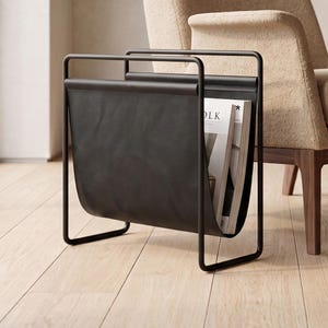 May include: A black metal magazine rack with a smooth, dark leather-like sling pocket. The rack features a rectangular frame design and holds a magazine with visible white pages and text, standing independently on a light-colored wooden floor.