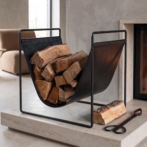 May include: A black metal log holder with a U-shaped sling design, filled with stacked brown firewood logs. A single log rests on the base next to the holder, accompanied by a pair of black metal fireplace tongs with long handles and looped grips.