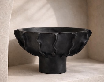 Könnte beinhalten: Eine schwarze dekorative Schale mit einem Sockel. Die Schale hat ein strukturiertes, wellenförmiges Design am Rand. Die Schale besteht aus einem dunklen Material und ist vor einem neutralen Hintergrund platziert.