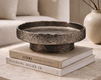 Könnte beinhalten: Ein dekoratives Tablett aus dunklem Metall mit strukturiertem Rand steht auf zwei Büchern. Die Bücher tragen die Titel "AT100: A Century of Style" und "Interiors Atelier AM". Im Hintergrund befindet sich eine Vase in neutralen Farbtönen.