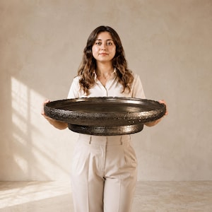 May include: A large, circular, dark-toned decorative tray with a textured, rugged outer rim and a smooth, flat interior surface. The tray features a raised, tiered base design with a matching dark, metallic-like finish and a wide, shallow profile.