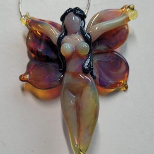 May include: A glass pendant necklace featuring a nude fairy design with outstretched wings. The fairy has black hair and a translucent body with yellow, orange, and purple hues. The wings are multi-colored with a glossy finish, and the necklace has a silver chain.