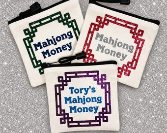 May include: Three off-white canvas zipper pouches with black trim. Two pouches feature a green or red glitter geometric border with the text Mahjong Money. The third pouch has a purple glitter border and the text Tory's Mahjong Money in blue.
