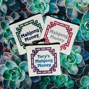 May include: Three white canvas zipper pouches with glittery text that reads "Mahjong Money" and "Tory's Mahjong Money". The text is framed by a decorative border. The pouches have black zippers and are set against a background of green succulents.