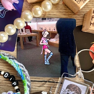 May include: A square image featuring a cartoon character in a pink outfit with cat ears and striped blue socks. The character is in an Apple store. The image is surrounded by jewelry, a keychain, and other small items on a wooden surface.