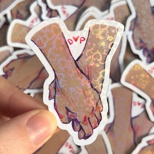 May include: A sticker featuring two hands clasped together, with a holographic overlay of hearts. The sticker has a white border and the letters "DVP" are visible. The overall color scheme is warm, with shades of brown and pink.