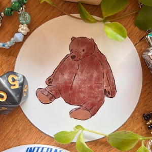 May include: A brown teddy bear illustration on a white circular surface. The bear is depicted in a seated position with its arms and legs extended. The artwork has a hand-drawn aesthetic. The image is surrounded by other objects.