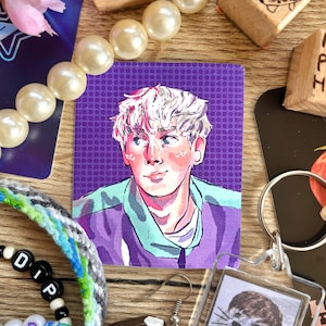 May include: A square sticker featuring a portrait of a person with white hair, wearing a purple jacket with a teal undershirt. The background is a purple dotted pattern. Other items include a pearl necklace, beaded bracelets, and a keychain.