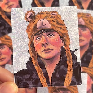 May include: A sticker featuring a portrait of a person wearing a brown bear hat. The portrait has a soft, painterly style with rosy cheeks and star-shaped accents. The background is a shimmering, glittery effect.