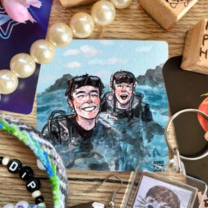 May include: A square watercolor painting depicts two people in scuba gear smiling in the ocean. The painting is surrounded by various accessories, including a pearl bracelet, beaded jewelry, and a keychain.