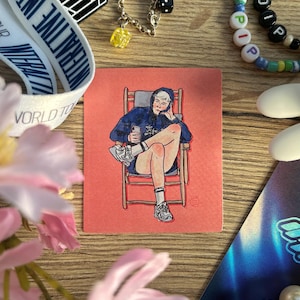 May include: A hand-drawn illustration of a person sitting in a director's chair. The person is wearing a blue hoodie, shorts, and white sneakers. The person is holding a cup in their hand. The illustration is on a pink background.