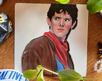 May include: A portrait of a man with dark hair, wearing a brown jacket and a red scarf, set against a neutral background. The artwork is on a small, rectangular card. The image is surrounded by various objects, including dice and a bracelet.