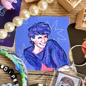 May include: A square sticker featuring a smiling person with blue hair and a blue shirt, set against a blue background with white starbursts. The sticker is surrounded by other items, including a pearl necklace, beaded bracelets, and a keychain.