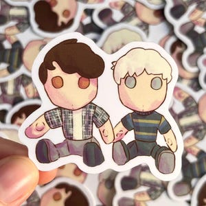 May include: A sticker featuring two cartoon figures holding hands. One figure has brown hair and wears a plaid shirt, the other has white hair and a striped shirt. Both figures have large, round eyes and are seated.