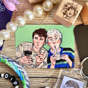 May include: A small, rectangular art print featuring two people, one holding a wine glass and the other a beverage in a glass with a straw. The print is on a light green background, surrounded by other small objects.