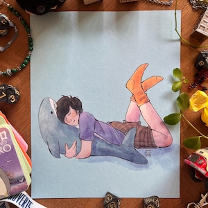 May include: Illustration of a person lying on a shark plushie, wearing a purple shirt and orange socks. The artwork is on a light blue background. Various dice and small wooden blocks are scattered around the artwork.