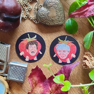 May include: Two round stickers with black backgrounds feature illustrations of people with their heads replaced by strawberries. One person is laughing and the other has a neutral expression.