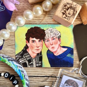 May include: A rectangular art print featuring two young men with detailed facial features. One has dark hair and a plaid shirt, the other has white hair and a blue sweater. The background is yellow. Other items include a stamp and a beaded bracelet.