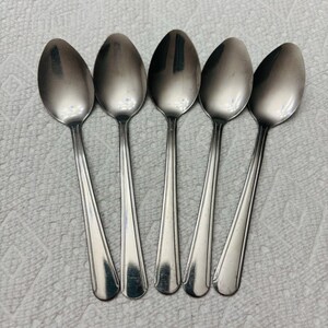 Puede incluir: Cinco cucharas de metal plateado dispuestas en fila. Cada cuchara presenta un cuenco ovalado liso y un mango delgado con un sutil detalle de línea vertical que recorre el centro, terminando en una base redondeada y ligeramente ensanchada.