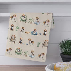 May include: A beige kitchen towel with a repeating pattern of beagle dogs in various poses, including sitting in armchairs and lying on cushions. The towel is hanging on a white shelf above a wooden countertop. The background includes a plant and a basket of bread.