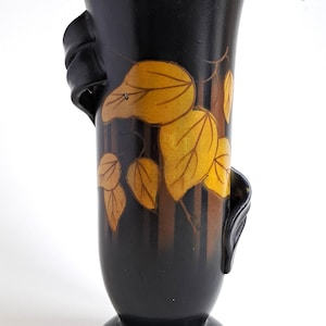 May include: Black ceramic vase with a flared top and a pedestal base. The vase features a design of yellow and brown leaves and vertical lines. Two curved handles are on the sides.