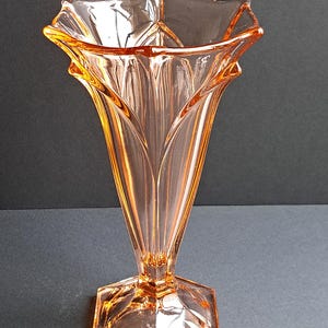 Può includere: Un vaso di vetro color pesca con base esagonale e parte superiore svasata a forma di stella. Il vaso ha un corpo sottile e scanalato ed è alto circa 20 cm. Il vetro ha una superficie liscia e riflettente e il design è di ispirazione Art Déco.