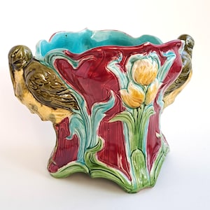 Könnte beinhalten: Ein Keramik-Pflanzgefäß mit einem lebendigen Design. Das Pflanzgefäß hat einen tiefroten Körper mit türkisfarbenen Akzenten und einem gelben Blumenmotiv. Zwei Vogelskulpturen sind in das Design integriert. Das Pflanzgefäß ist ein dekoratives Stück für die Inneneinrichtung.