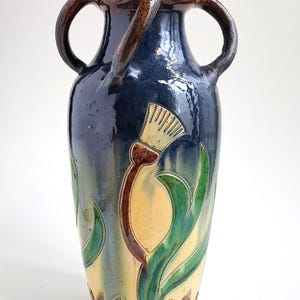 Pode incluir: Um vaso de cerâmica alto, com um esmalte azul escuro e duas alças arqueadas. O vaso apresenta um desenho floral em amarelo, verde e castanho. O vaso tem cerca de 30 cm de altura.