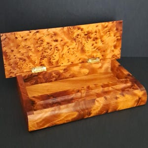 Puede incluir: Caja de madera abierta con tapa con bisagras. La caja y la tapa están hechas de madera de tonos cálidos con un patrón de vetas único y moteado. La caja es rectangular con bordes redondeados y un pequeño compartimento interior.