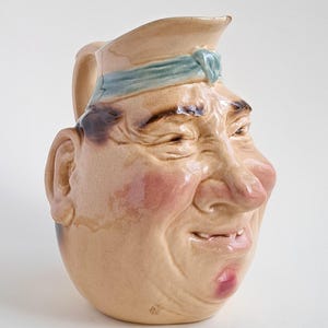 May include: A vintage ceramic pitcher shaped like a smiling man's face. The pitcher has a tan base color with pink cheeks and lips, and a blue band around the hat. The handle is on the left side, and the pitcher has a glossy finish.